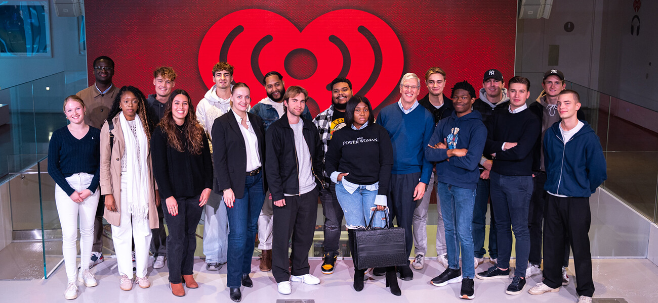 A diverse group of individuals stands together, smiling against a vibrant red iHeartRadio background. Stylish outfits showcase personal flair