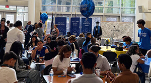 A vibrant college event with students at tables, balloons, banners, and informational displays in a bustling indoor setting.
